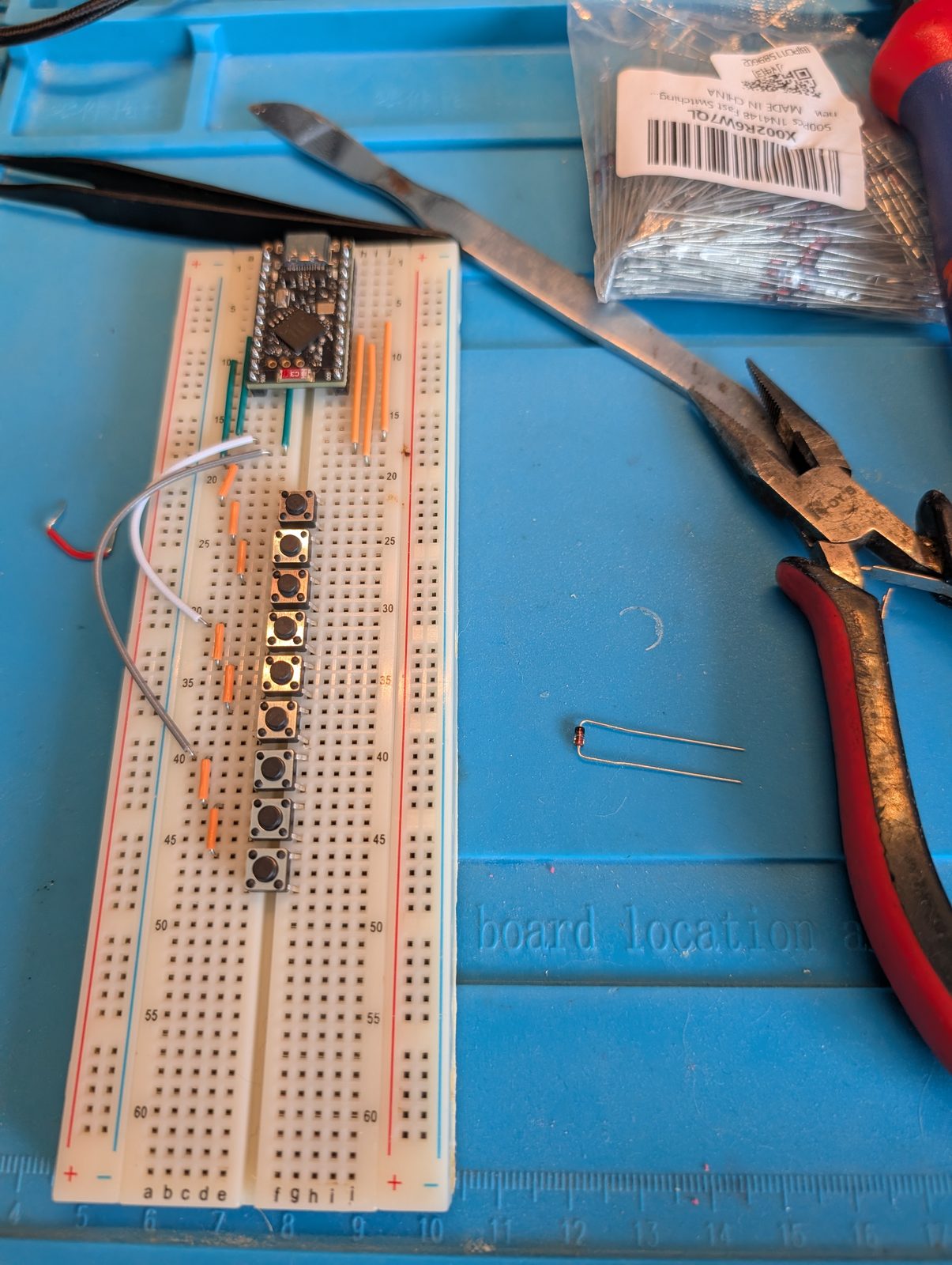 A breadboard on a blue work mat with a nice!nano at the top and a column of eight tactile switches wired below it, next to a bag of 1N4148 diodes, tweezers and pliers.