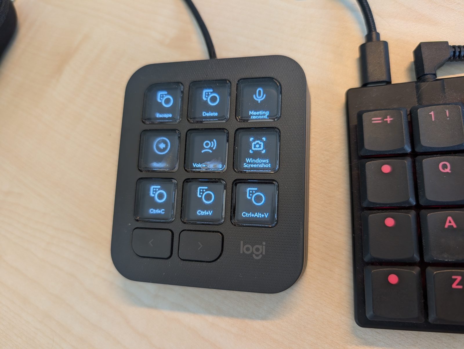 The Logitech MX Creative Console on a desk beside a mechanical keyboard, its nine keys lit with programmed labels — Escape, Delete, Meeting record, Windows Screenshot, Ctrl+C, Ctrl+V and Ctrl+Alt+V — with the USB-C cable running off the top of the frame.